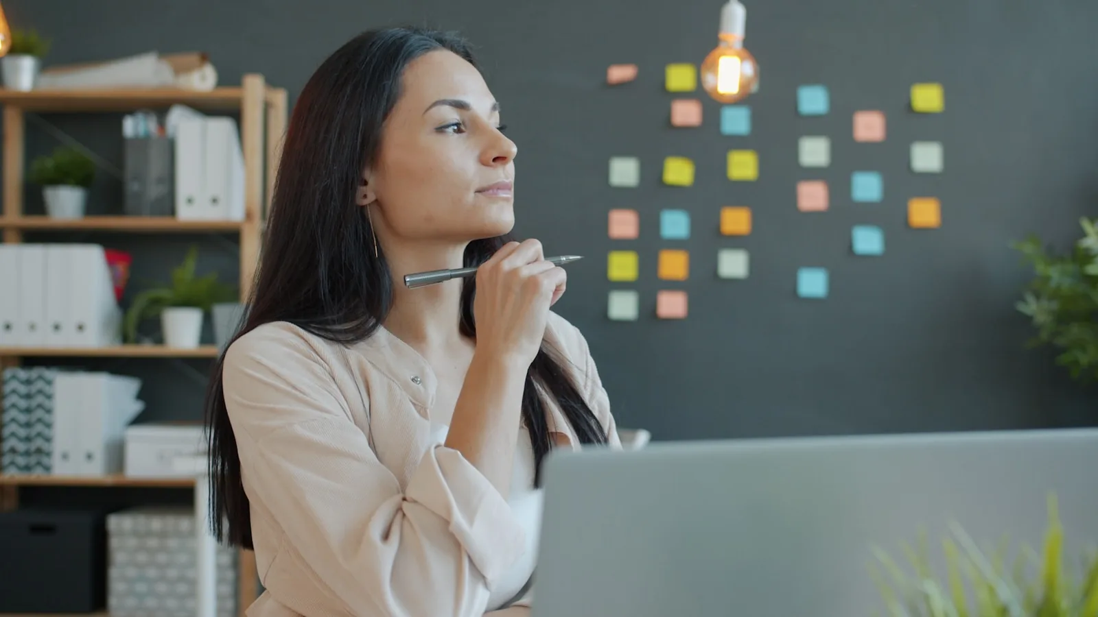 Entrepreneure pensive à son bureau, stylo en main, devant un mur de post-it d'organisation
